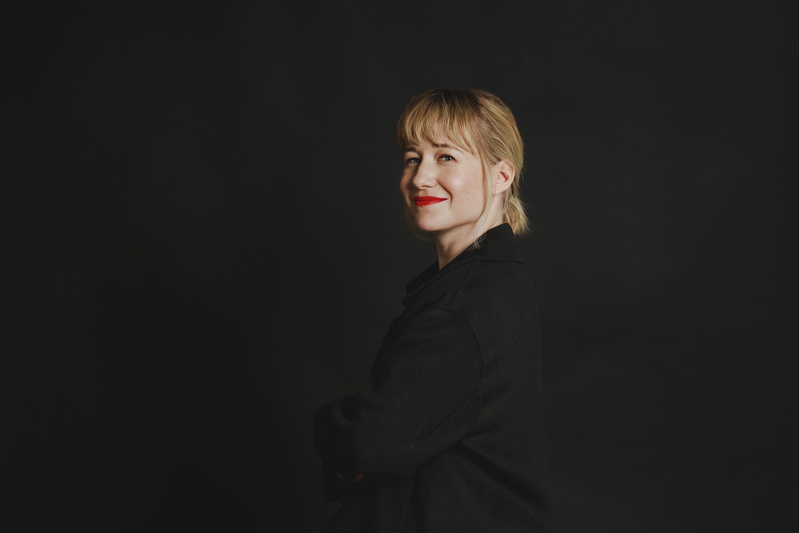 Studio portrait of Linda Liukas in a dark jacket, arms folded, turned towards the camera against a dark background.